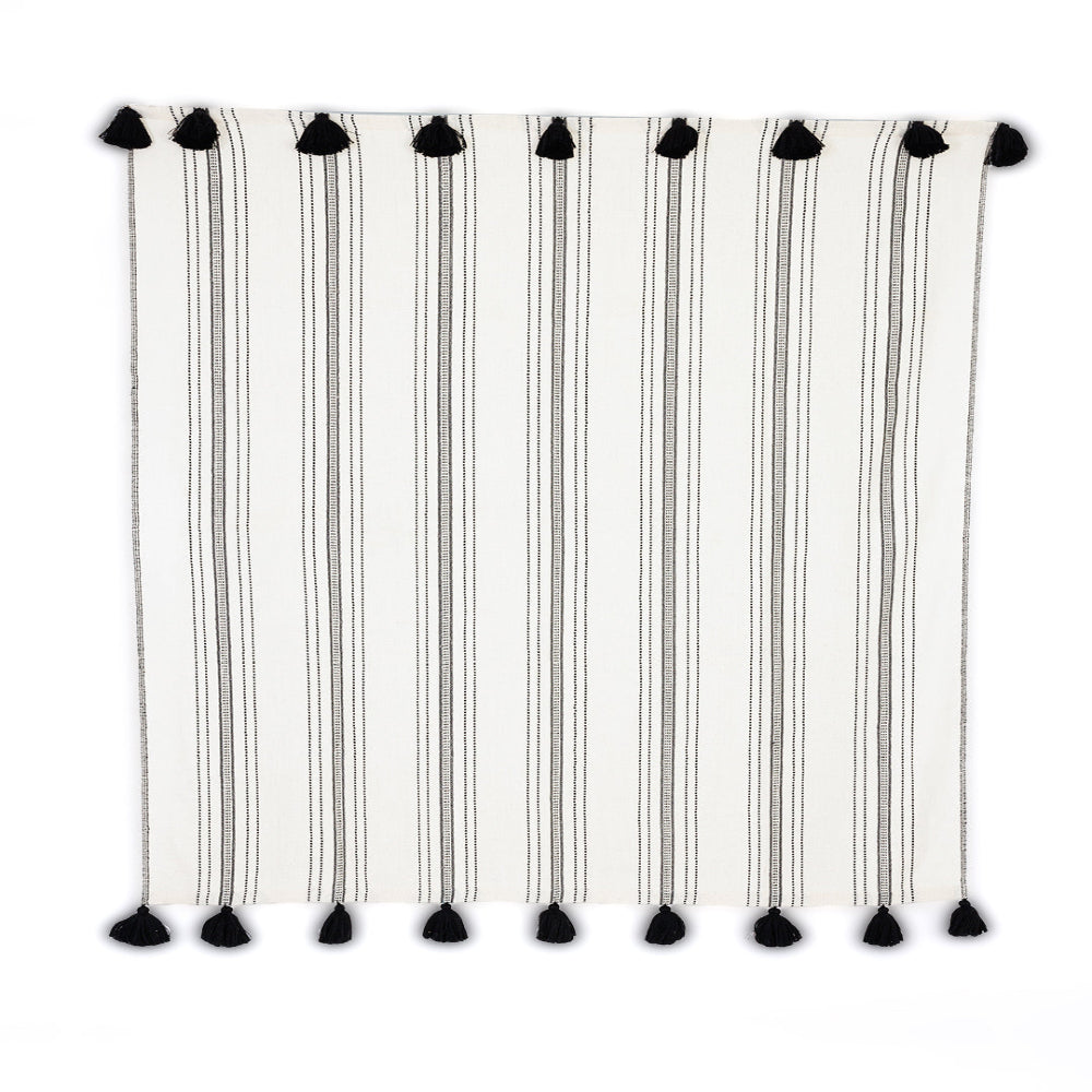 A striped throw blanket with ivory and ink colors, featuring handmade black pom poms on both ends, displayed on a plain background.