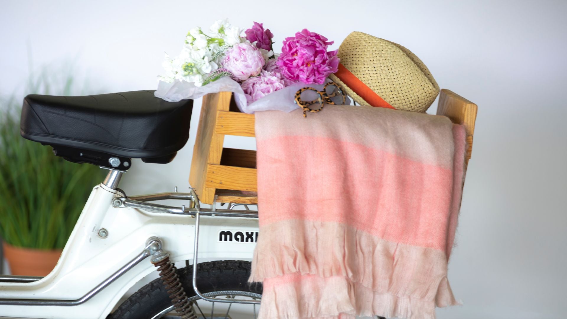 Rear view of white bicycle with crate carrying pink blooms, straw hat, and striped blanket evoking heritage, craftsmanship, and lifestyle charm