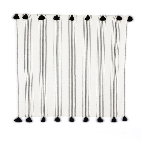 A striped throw blanket with ivory and ink colors, featuring handmade black pom poms on both ends, displayed on a plain background.