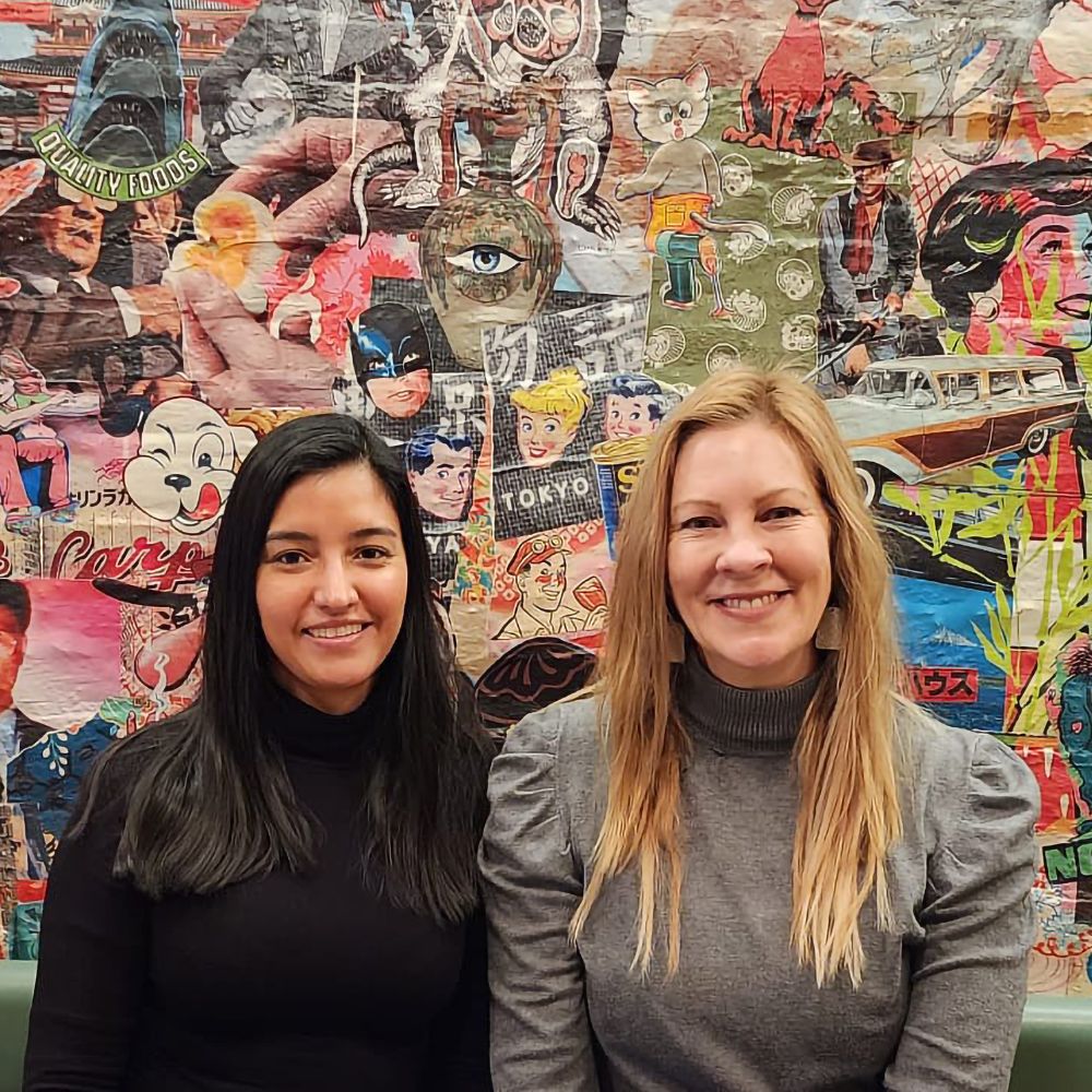 Two women pose in front of a vibrant poster wall, showcasing a variety of artistic designs and messages.