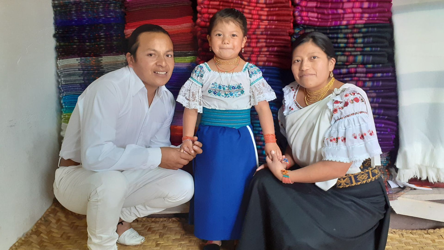 Three individuals in traditional embroidered attire seated before vibrant folded textiles celebrating Palestinian heritage and artisanal craftsmanship